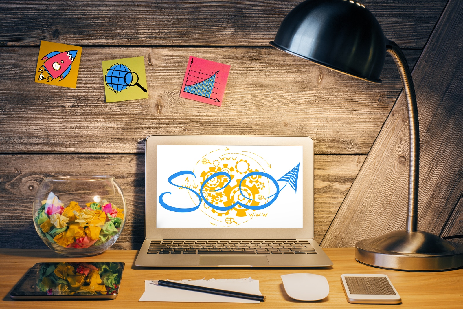 Creative designer desktop with table lamp SEO sketch on laptop scree various devices and stickers with rocket icons on wooden wall. SEO and internet concept