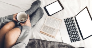 Image of a woman sitting on her bed with a book, tablet, and laptop out. She is reading her therapists blog. Her therapist worked on their private practice SEO using blogging techniques.