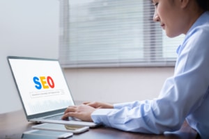 A woman smiles as she types on a laptop with the text SEO on the screen. We offer support for private practice website. Contact us to learn how we support local SEO for therapists today. 