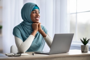 Happy black muslim woman at a computer staring out the window. She is working on SEO and writing copy for SEO after reading a blog by an SEO specialist at Simplified SEO Consulting