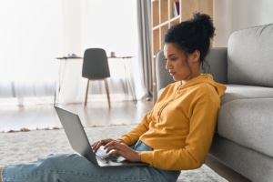 young woman sits in her living room typing on her computer as she blogs for SEO. Learn SEO and more about copywriting from an SEO expert at Simplified SEO Consulting
