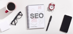 Minimalist photo of a notebook that says "SEO" with a couple other business tools including an alarm clock, a phone, classes and a cup. Representing how our team helps you get to the top of Google by simplifying the SEO process.