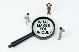 Photo of toy figures staring at the words "what makes you unique" through a magnifying glass. This photo represents how adding your unique approach to therapy can help you when using your website to market to a counseling niche.