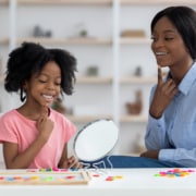 Photo of a female, black speech therapist helping a young girl with articulation using a mirror. This represents the important work SLPs in private practice do. SEO can help get your services out to move potential patients.