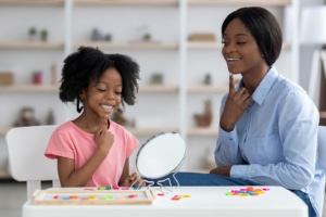 Photo of a female, black speech therapist helping a young girl with articulation using a mirror. This represents the important work SLPs in private practice do. SEO can help get your services out to move potential patients. Voice Search is the future of search, so it's important to consider optimizing for voice search as well.