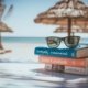 Shows a beach scene with a stack of books that have sunglasses on them. Represents how seo for mental health can help therapists with the summer slump.