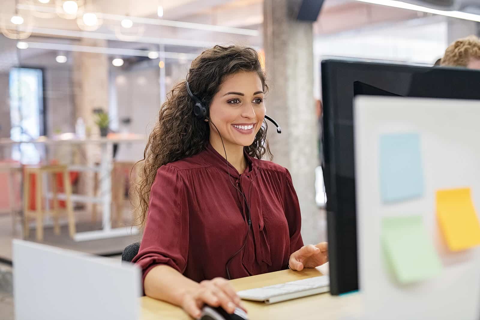 A woman working on a computer with a headset on smiling. Want to learn about SEO and how it impacts a business. We can teach you about SEO and it being an investment to you. 