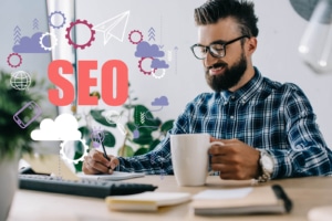 A man smiles while looking at his laptop and coffee cup. This could represent learning how to build seo for therapists. Learn to support your private practice seo today.
