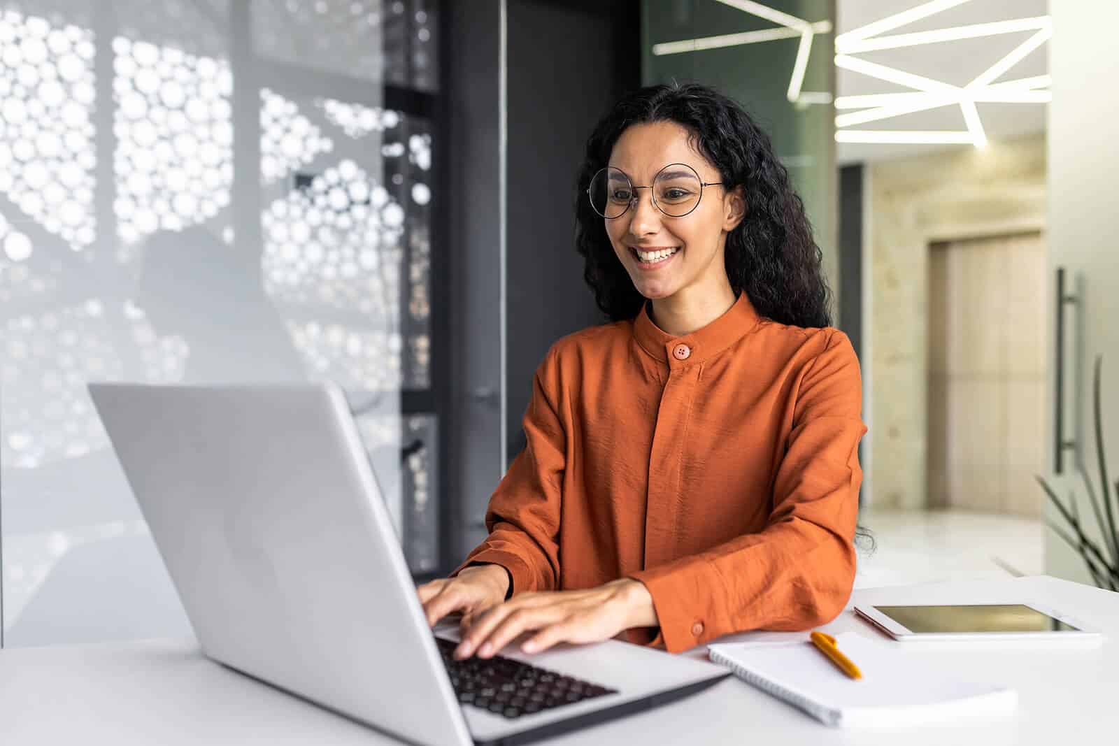 Image of a smiling woman wearing an orange shirt working on a laptop. This photo represents how you can maximize your online presence with the help of SEO.