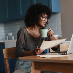 Shows a black, female therapist holding a cup of coffee and paper while smiling at her computer. Represents how online therapy advertising can be made easier with seo for therapists and seo keywords for mental health.