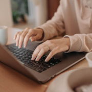 Shows a young woman wearing a pink sweatshirt typing on her laptop to create service page content. Represents how counselor seo and search engine optimization for counselors can boost your practice's rankings.