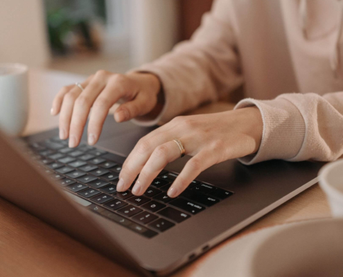 Shows a young woman wearing a pink sweatshirt typing on her laptop to create service page content. Represents how counselor seo and search engine optimization for counselors can boost your practice's rankings.