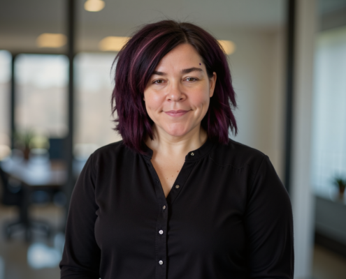 Mary Walker – Co-owner of Simplified SEO Consulting, with shoulder-length dark purple hair wearing a black button-up shirt, smiling slightly, standing in a bright office space with blurred windows in the background