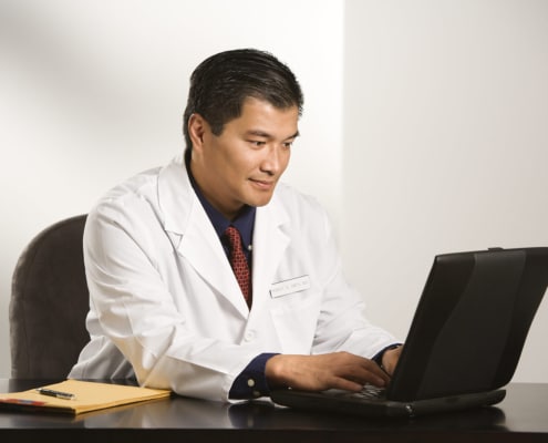 An Asian american male doctor typing on a black laptop at his desk. Our SEO for functional medicine doctors is here to help you wellness website rank higher on Google. Find your ideal clients today with our SEO services.
