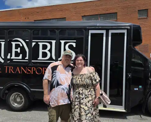 Mary and Edward Walker – Co-owners of Simplified SEO Consulting, standing together smiling in front of a black shuttle bus on a sunny day. Edward wears a palm-leaf patterned shirt and khaki shorts, and Mary wears a floral dress and black sandals.