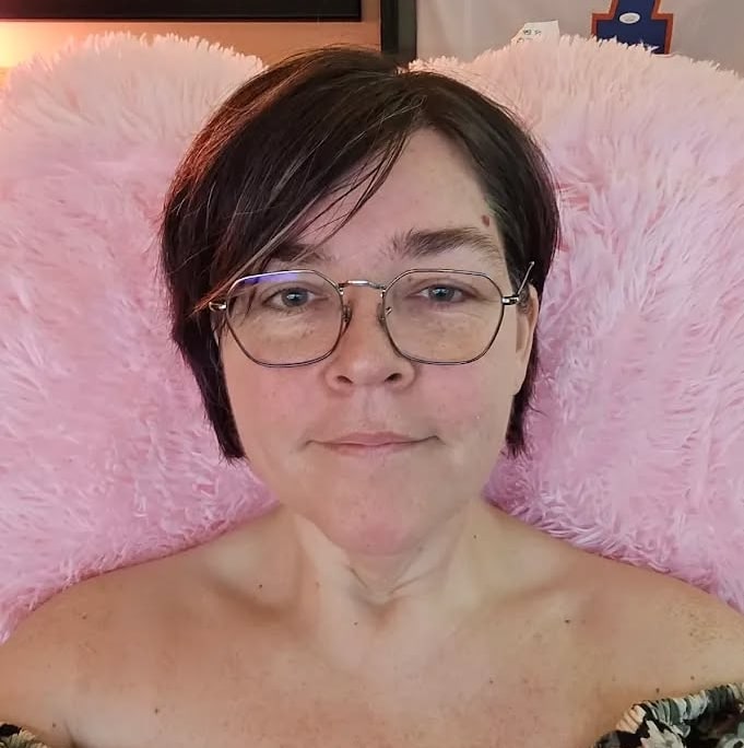 Mary Walker – Co-owner of Simplified SEO Consulting, with short dark hair wearing glasses and an off-the-shoulder patterned top, sitting in a fluffy pink chair with sports jerseys framed on the wall behind her.