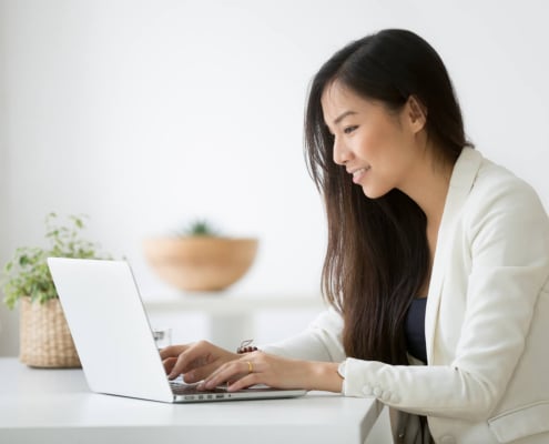 Happy woman sitting at a desk smiling while typing on a laptop. Learn to improve your private practice SEO and begin to find your ideal clients with the support of Simplified SEO Consulting.