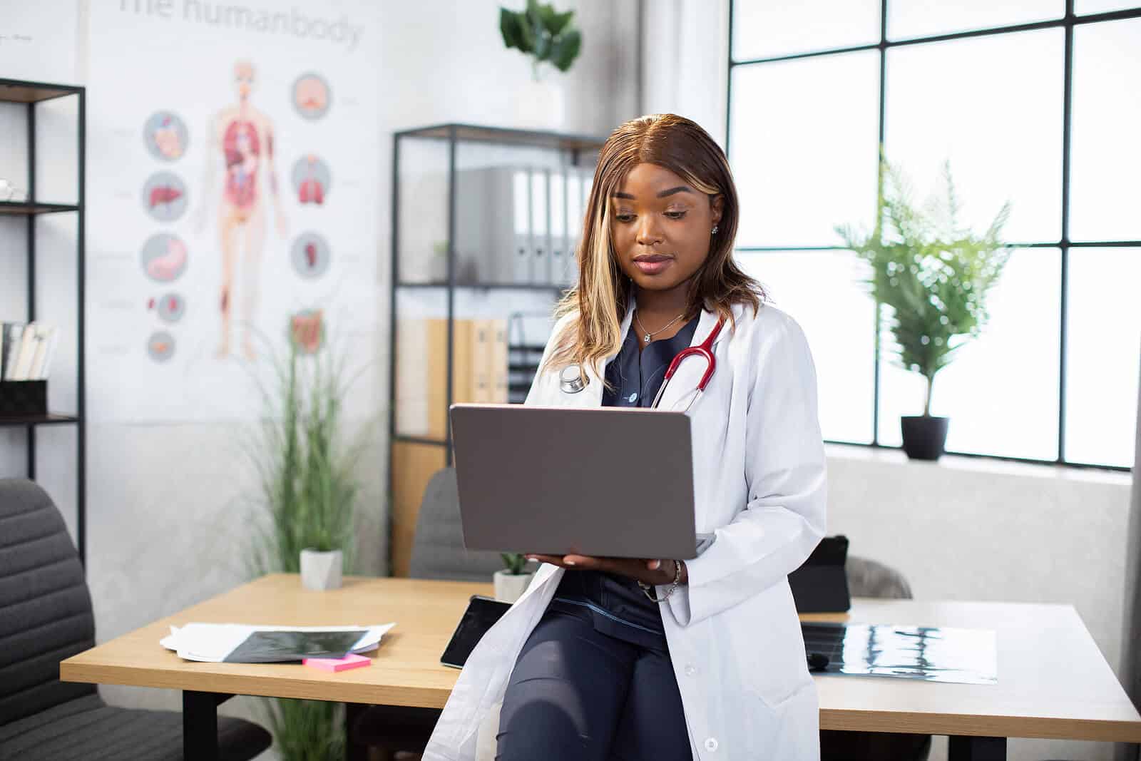 Thoughtful woman wearing a doctors white coat leaning against a desk holding a laptop. Begin reaching people who truly benefit from your holistic approach to health with functional medicine SEO. 