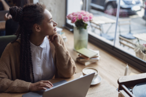 woman looking out window | social media company | social media marketing agency | social media marketing | Tampa, FL | Miami, FL | Bradenton, FL A biracial woman sitting near a window working on her laptop as she looks curiously. If you're a professional wondering if you should work on SEO or social media, we're here to help. Read our blog for more info!