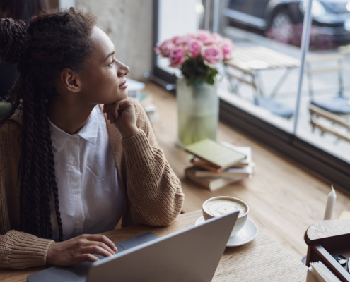 A biracial woman sitting near a window working on her laptop as she looks curiously. If you're a professional wondering if you should work on SEO or social media, we're here to help. Read our blog for more info!