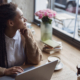 A biracial woman sitting near a window working on her laptop as she looks curiously. If you're a professional wondering if you should work on SEO or social media, we're here to help. Read our blog for more info!