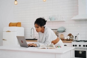 Young woman working on a laptop in a bright, modern kitchen surrounded by books and highlighters, showcasing the importance of therapy SEO and health keywords in creating engaging content for alternative healing practitioners to boost online visibility.