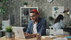 Young man in a red beanie typing on a laptop at a wooden desk with design tools and plants, showcasing the benefits of simplified SEO and SEO for coaches in building engaging content for alternative healing practitioners.
