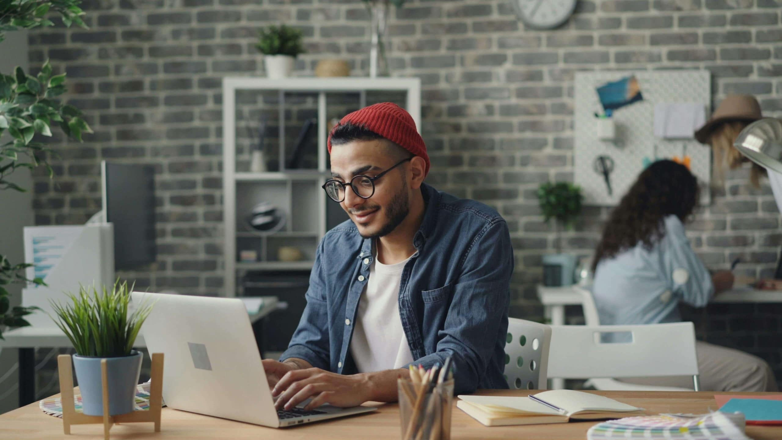 Young man in a red beanie typing on a laptop at a wooden desk with design tools and plants, showcasing the benefits of simplified SEO and SEO for therapist in building engaging content for therapy practices.