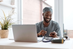 Smiling medical professional in grey scrubs using a smartphone at a desk with a laptop and stethoscope, showcasing the value of SEO consulting services and alternative healing SEO in enhancing online presence for healthcare practitioners.