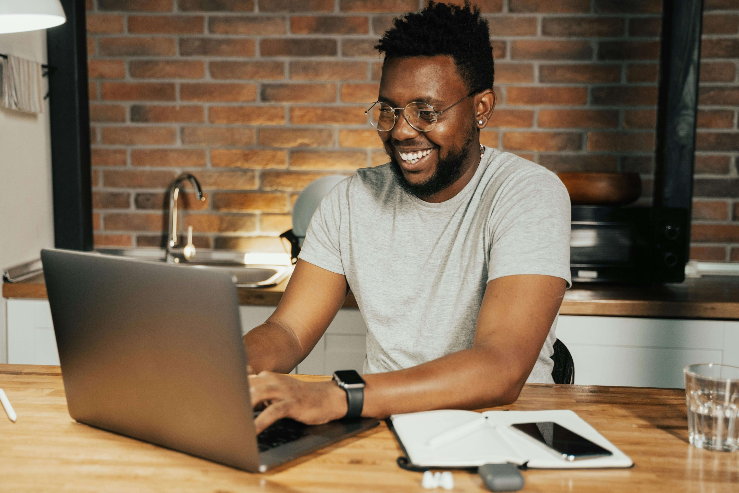 Smiling man wearing glasses working on a laptop at a kitchen table with a notebook and phone nearby. Integrative medicine SEO helps practice owners create content that connects their expertise to the patients actively searching for holistic care.