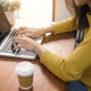 Woman in yellow sweater typing on laptop at wooden desk with coffee cup. Discover how SEO for online therapy services can bring clients directly to your practice.