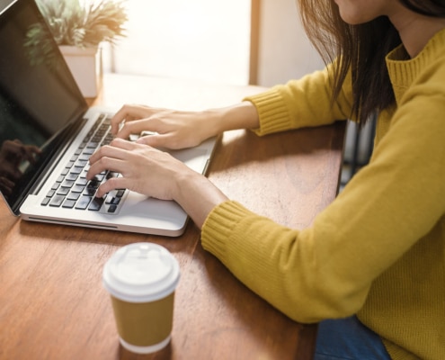 Woman in yellow sweater typing on laptop at wooden desk with coffee cup. Discover how SEO for online therapy services can bring clients directly to your practice.