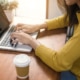 Woman in yellow sweater typing on laptop at wooden desk with coffee cup. Discover how SEO for online therapy services can bring clients directly to your practice.