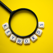 White letter tiles spelling "KEYWORDS" arc around the bottom of a black magnifying glass on a yellow background. Are you searching for the right keywords to make your online counseling pages stand out in search results? SEO services for therapists can help you find and use them strategically.