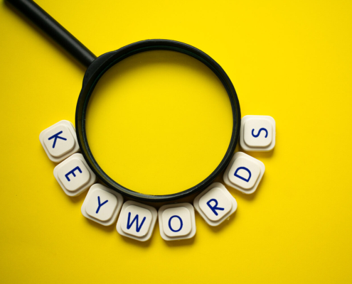 White letter tiles spelling "KEYWORDS" arc around the bottom of a black magnifying glass on a yellow background. Are you searching for the right keywords to make your online counseling pages stand out in search results? SEO services for therapists can help you find and use them strategically.