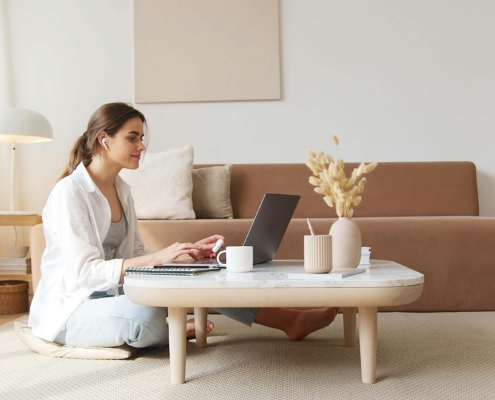 Woman sitting on the floor working on a laptop at a coffee table in a cozy living room, representing how SEO consultants and SEO consulting services help therapists create authentic content for their practice from anywhere.