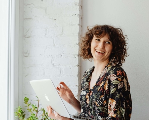 A woman with curly hair smiles while holding a laptop near a bright window with a small potted plant beside her. SEO for therapists makes it possible to spend less time searching for clients and more time doing the work you love.