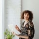 A woman with curly hair smiles while holding a laptop near a bright window with a small potted plant beside her. SEO for therapists makes it possible to spend less time searching for clients and more time doing the work you love.