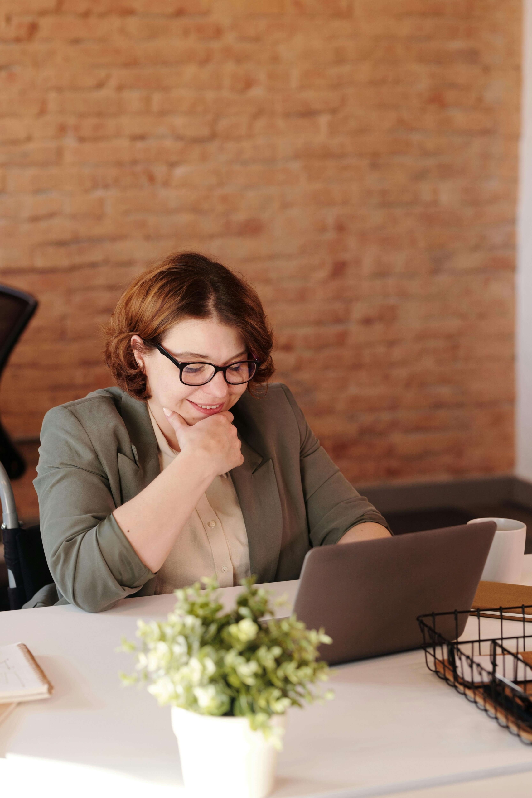 A woman with short red hair and glasses smiling while working on a laptop at a desk with a small plant nearby. Finally feeling confident online is possible. Discover what SEO help for therapists looks like at Simplified SEO Consulting.