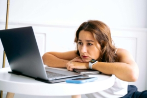 A young adult woman sitting staring blankly at her laptop screen. Representing how social media can feel overwhelming to people. Discover how our social media management package can help.