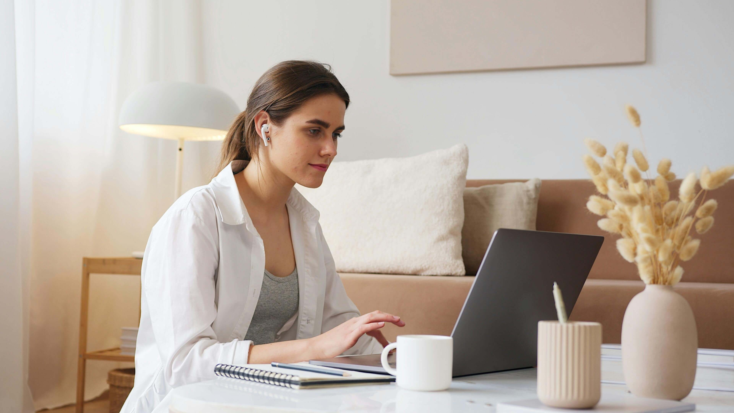 woman using laptop | keywords for online counseling pages | seo services for therapists | seo company for counselors | Michigan | Missouri | Wyoming A woman with a ponytail wearing a white shirt sits at a desk with a laptop, notebook, and coffee mug in a bright neutral toned living space with wireless earbuds in. SEO for private practices works quietly in the background so you can stay focused on what you do best, showing up for your clients.