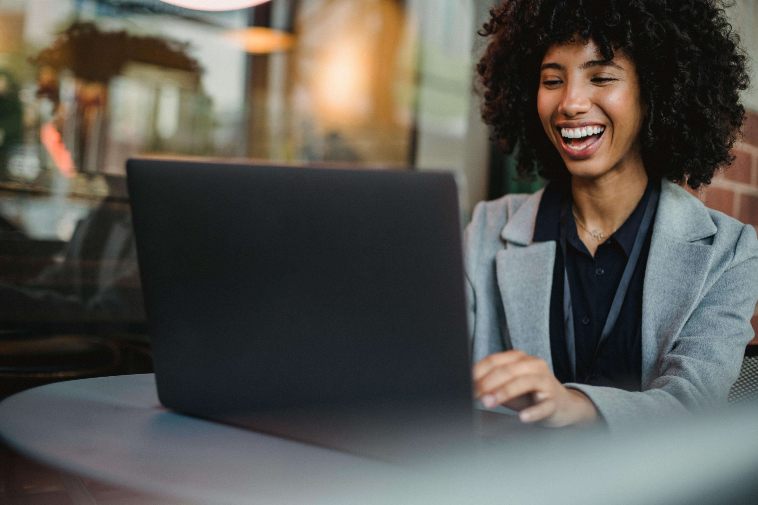 Woman smiling while sitting at a table using her laptop, symbolizing connection and client outreach through SEO for online therapy services.