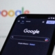 Close-up of a phone showing the Google homepage with the search bar visible, with a computer screen displaying the same page in the background, representing SEO for online therapy services.