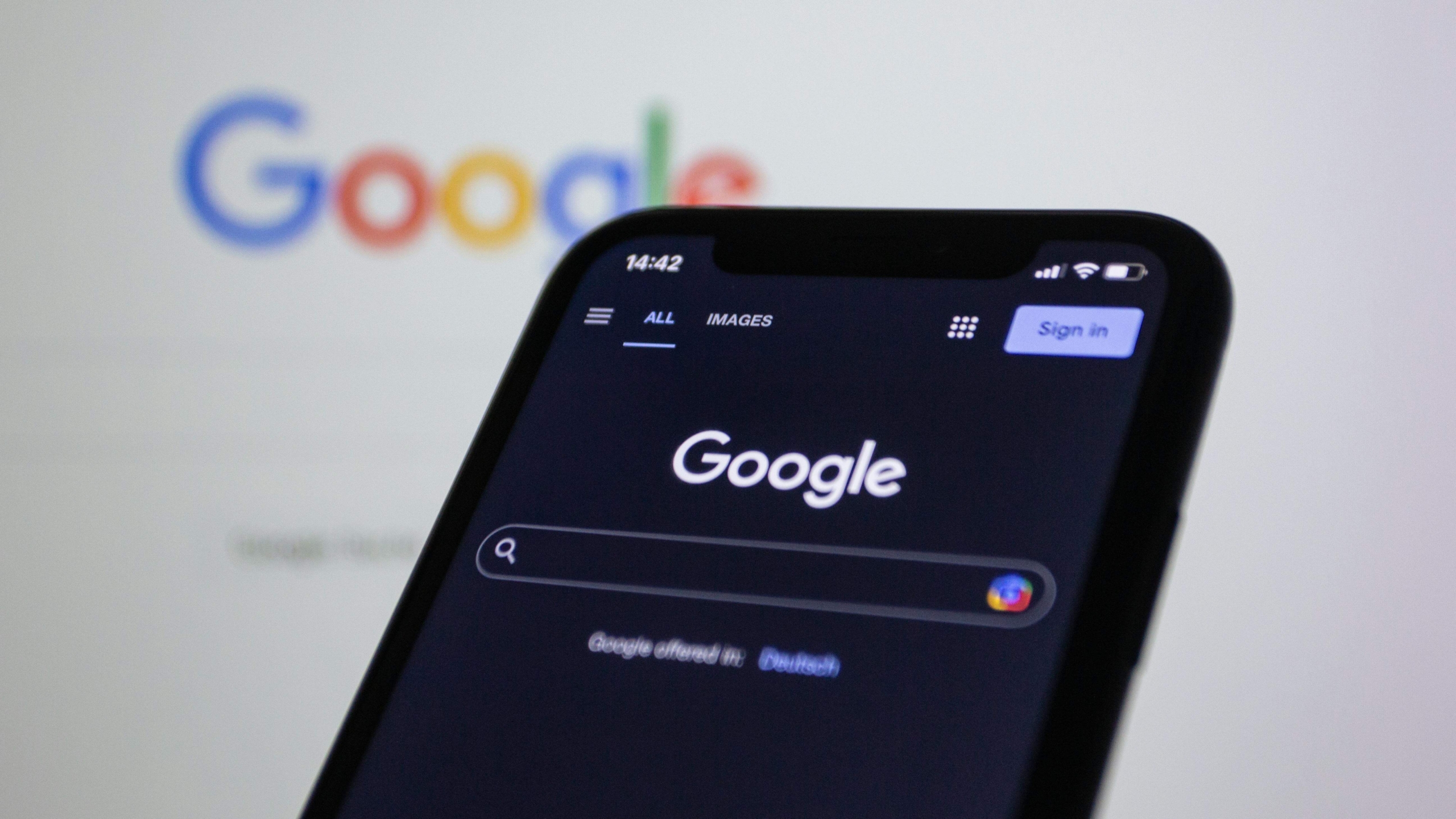 Close-up of a phone showing the Google homepage with the search bar visible, with a computer screen displaying the same page in the background, representing SEO for online therapy services.