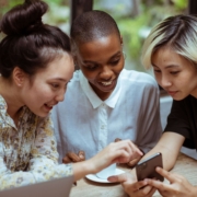 Three diverse women sitting together in a circle pointing at a phone. Representing social media marketing & sharing posts with friends. Discover how social media marketing can help you reach ideal therapy clients.