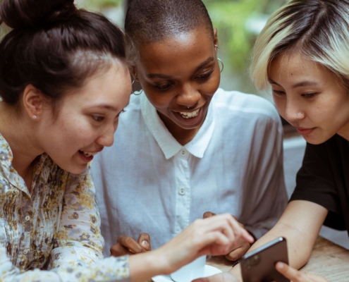 Three diverse women sitting together in a circle pointing at a phone. Representing social media marketing & sharing posts with friends. Discover how social media marketing can help you reach ideal therapy clients.