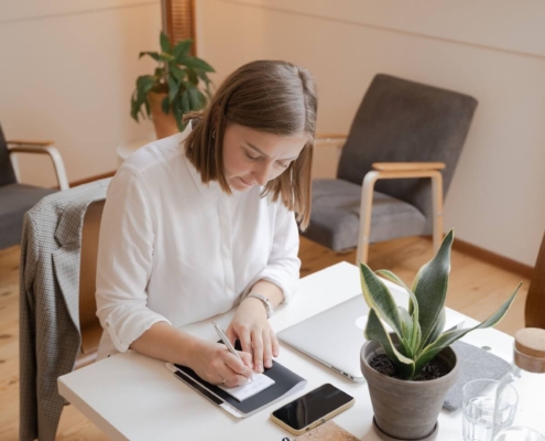 Woman writing in a notebook at a tidy desk with a laptop and plant nearby, representing how SEO consultants help with SEO for therapists by planning and refreshing content during the summer slowdown."