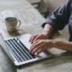 Close-up of a person working on a laptop with a coffee mug nearby, representing content creation and consistency in SEO help for therapists.