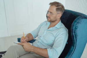 Man with glasses sitting in a blue armchair taking notes on a notepad, representing how therapists use seo marketing for therapists and the right keywords for counseling website content to plan ahead during the summer slowdown.