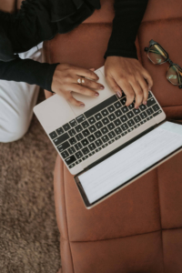 Overhead view of a person's hands typing on a laptop resting on a leather chair, with glasses nearby, representing how AI content marketing requires careful attention to AI accuracy before content goes live.